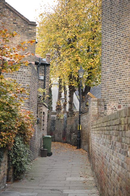 A narrow street in an urban residential area with tall, closely spaced brick buildings on either side. The buildings feature small windows, some with metal bars or shutters, and various external fixtu