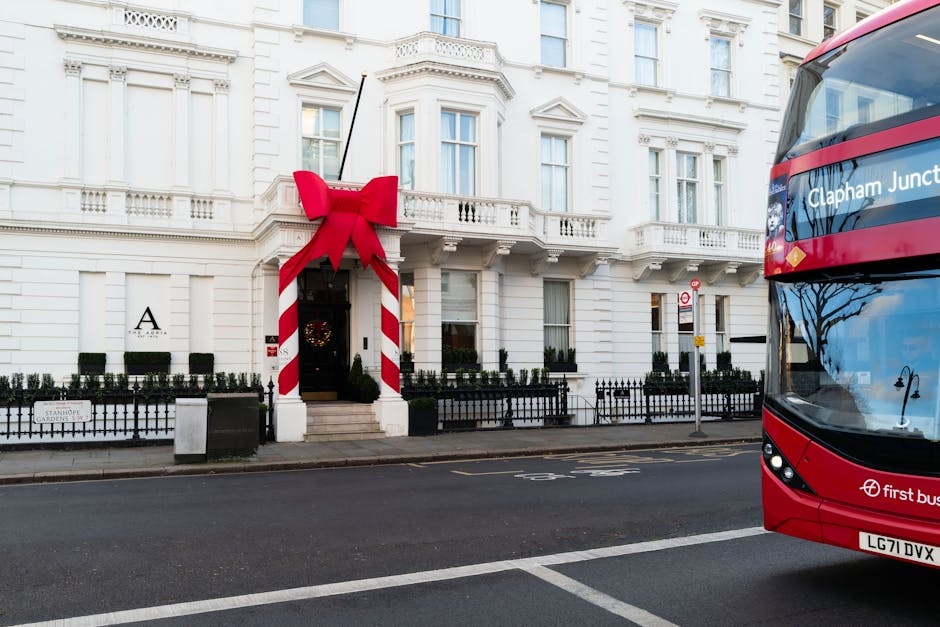 A white, multi-storey Victorian-style building with decorative architectural details and large windows, decorated for the holiday season with a large red ribbon and bow draped around the entrance archway, indicative of festive or special occasion preparations. In front of the building is a black metal fence and a small garden area with neatly trimmed bushes, and a sidewalk with a trash bin placed near the entrance. To the right, a red double-decker bus with the destination sign 'Clapham Junction' is partly visible, parked on the street which has marked lanes and road signs. The scene is well-lit with natural light, showcasing the street environment typical of Clapham, Lambeth, with a focus on the building's facade and the bus, suggesting urban residential or commercial premises involved in a home relocation or moving process, similar to services offered by Removal Company Lambeth.