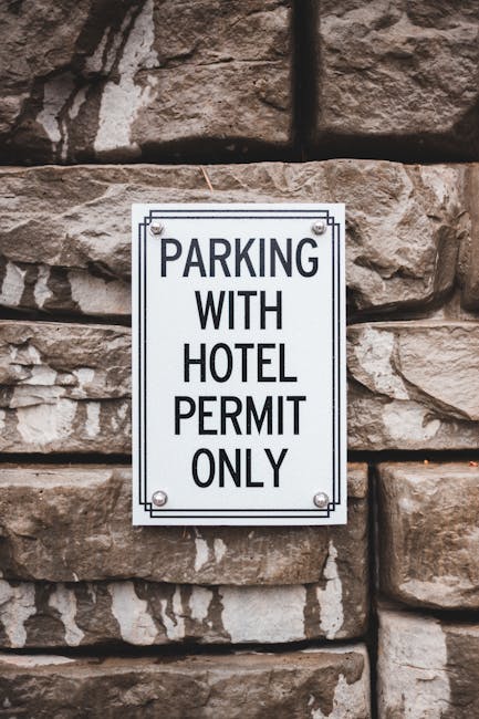 A rectangular metal sign affixed to a stone wall, displaying the message 'Parking with hotel permit only' in black uppercase letters on a white background. The sign is attached with four screws, one a